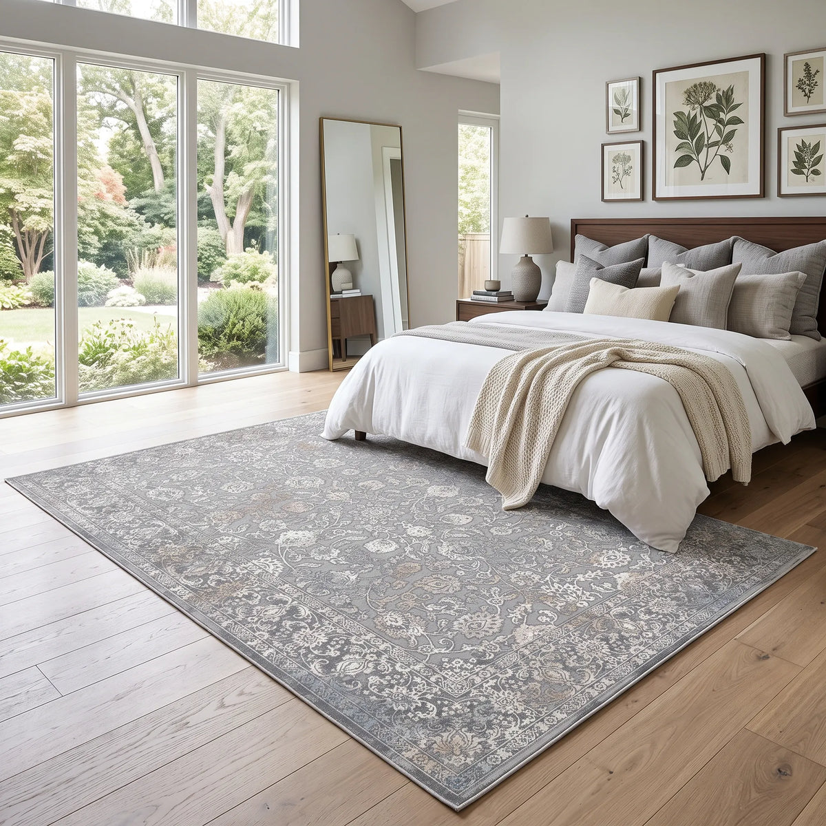 Un grande tappeto rettangolare con un intricato motivo floreale dall’effetto vissuto, nei toni del grigio, beige e bianco, disposto su un pavimento in legno chiaro in una camera da letto moderna.