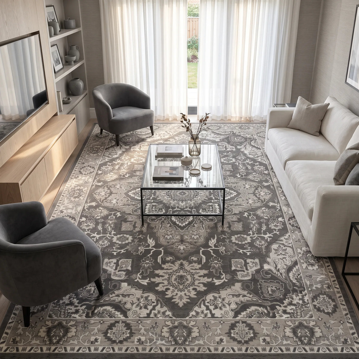 Un grande tappeto in stile tradizionale nei toni del grigio e del bianco sporco, inserito in un soggiorno moderno con un tavolino da caffè con piano in vetro, un divano grigio chiaro, due poltrone grigie e un mobile porta TV in legno.