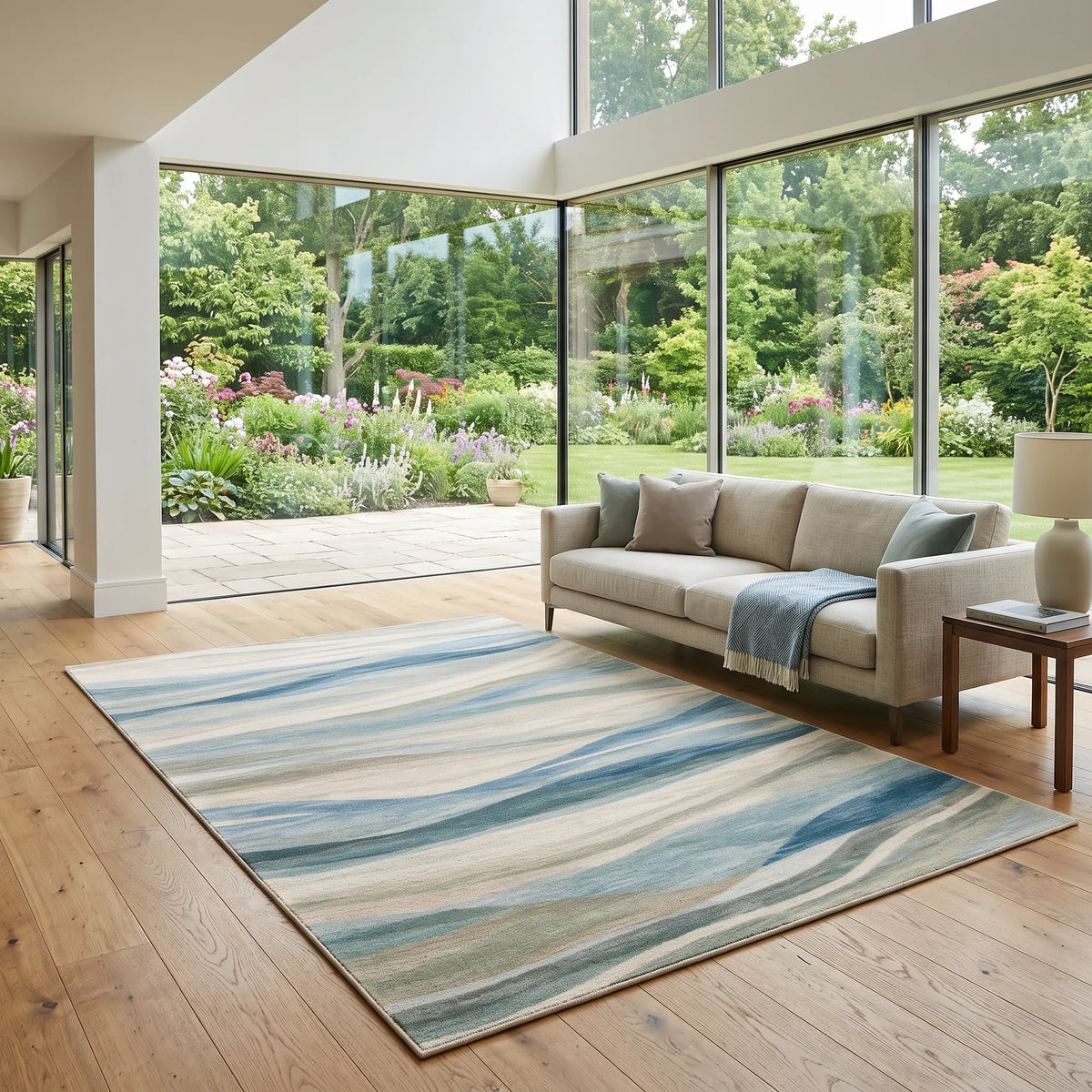 Un grande tappeto rettangolare con un motivo astratto ondulato nei toni del blu, del beige e del grigio, inserito in un moderno soggiorno con un divano beige chiaro e un pavimento in legno, affacciato su un rigoglioso giardino verde visibile attraverso ampie finestre a tutta altezza.